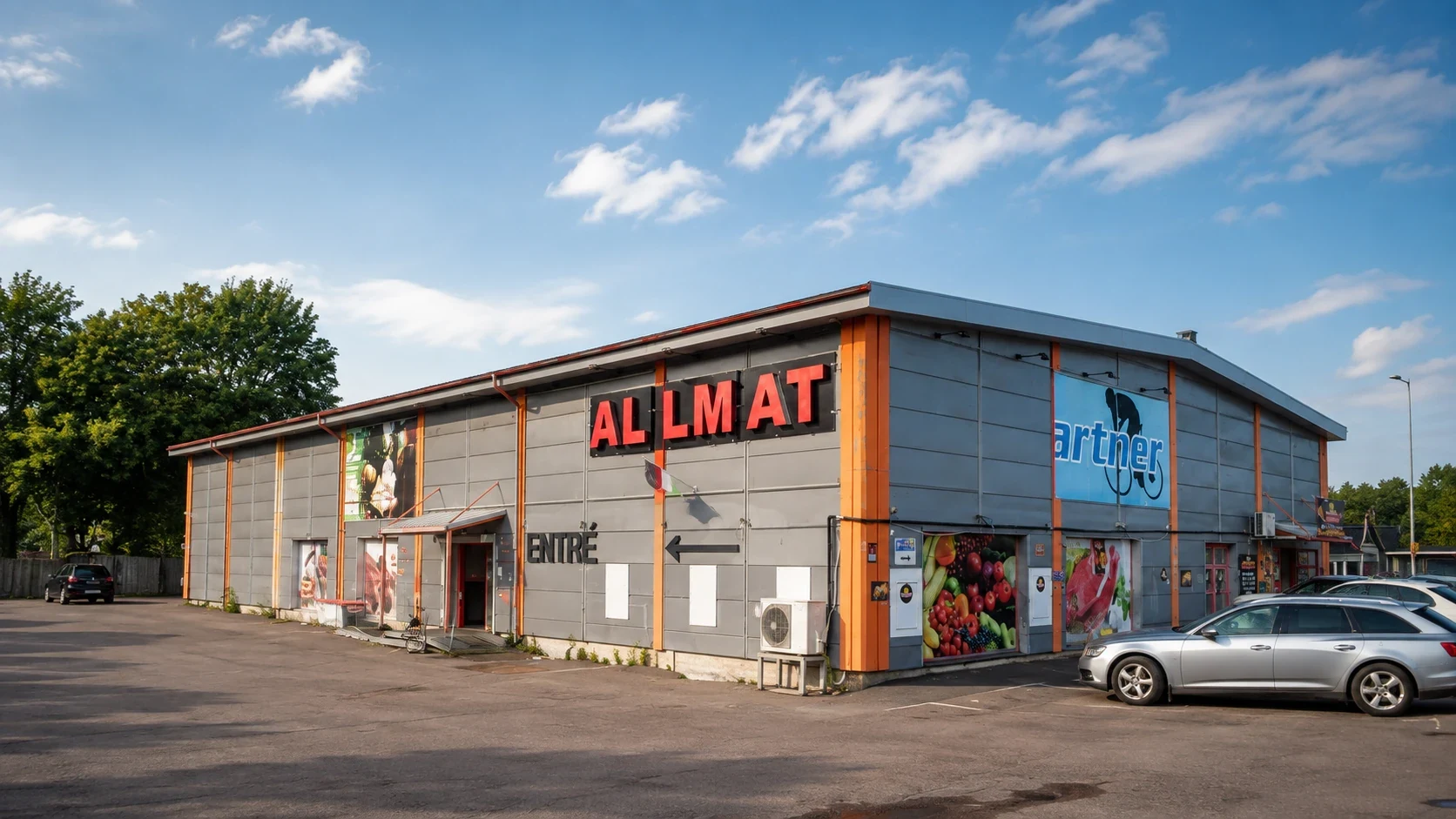 Allmats grå plåtfasad med orange pelare och röda skyltbokstäver, sedd från asfaltsparkeringen en solig dag.